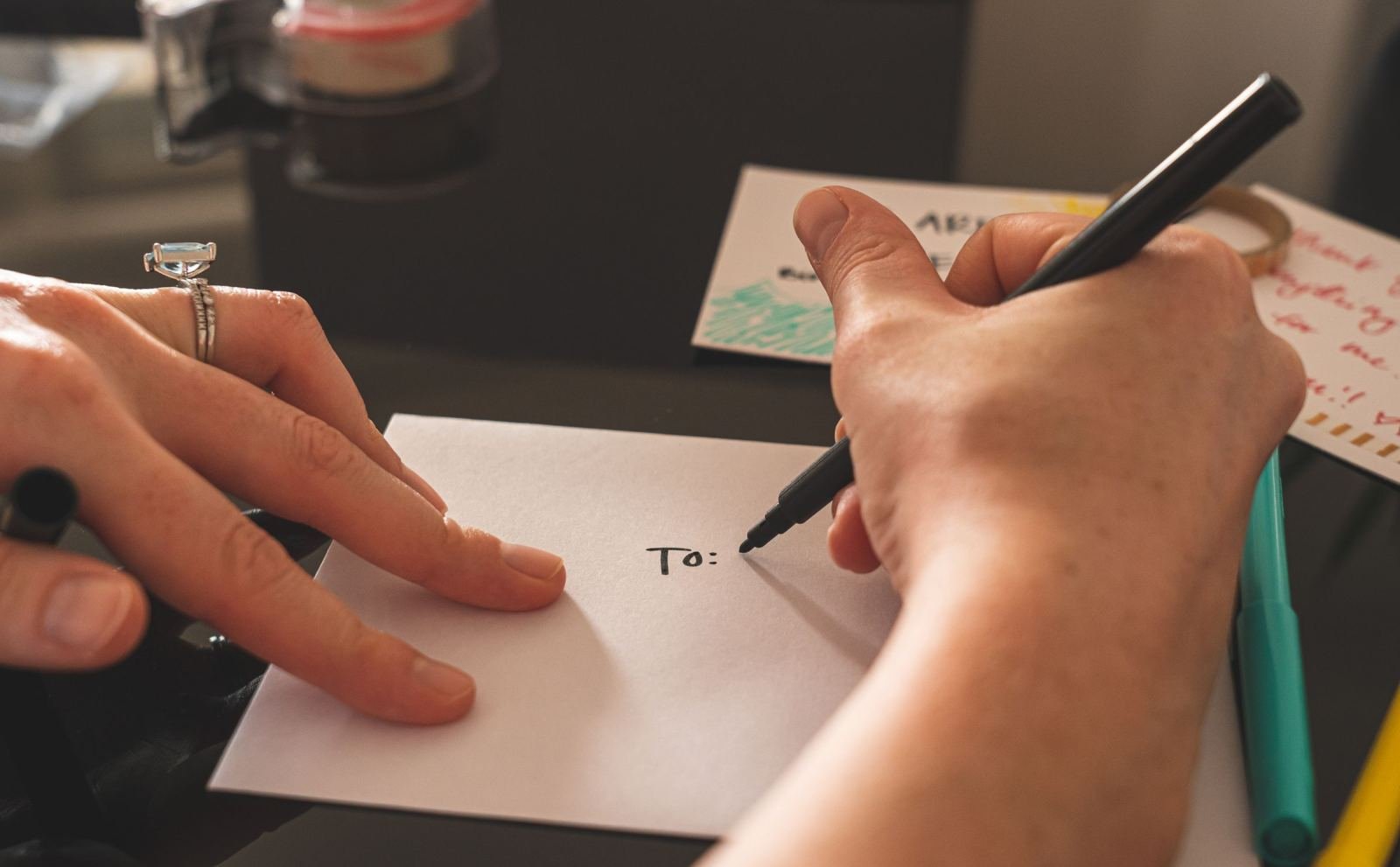 Two people's hands on a table as one writes 'To:' on a white card with a black marker