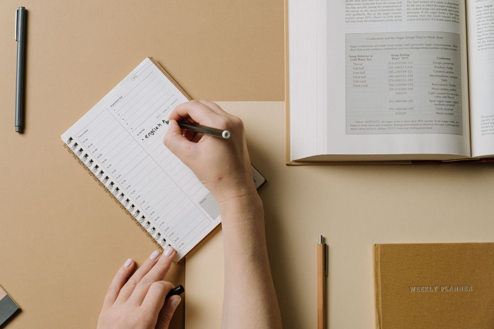 A person writing in a spiral notebook next to a textbook and a weekly planner