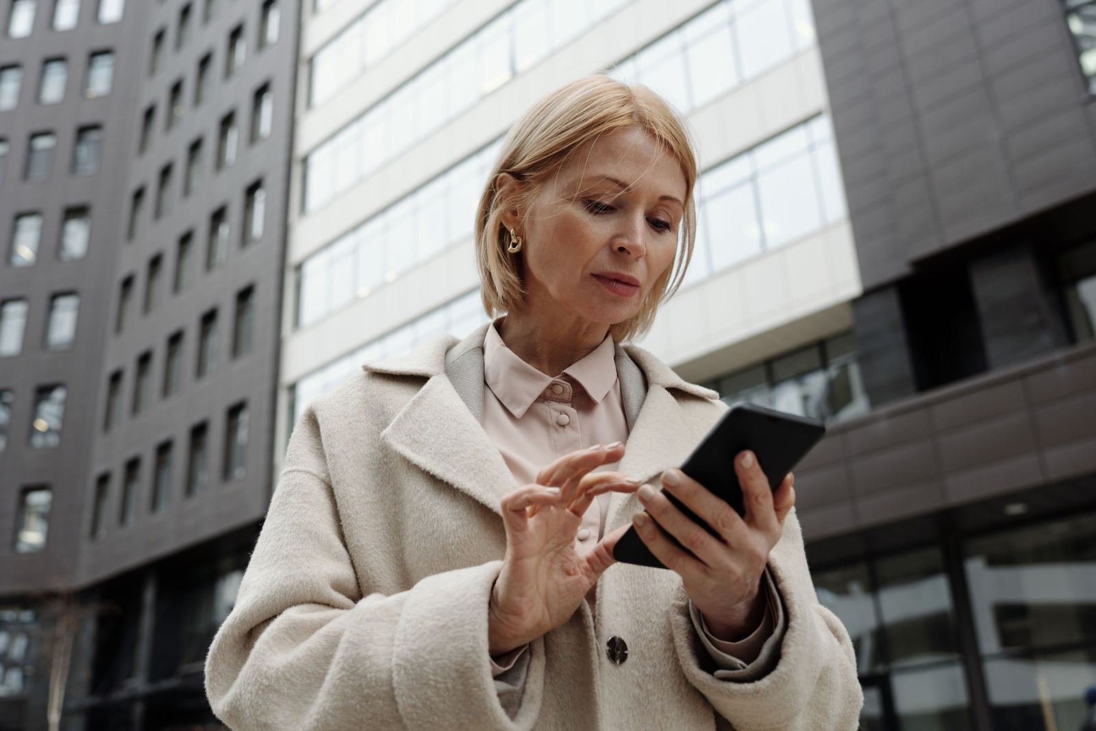 A woman in a beige coat looks at her smartphone in front of modern buildings
