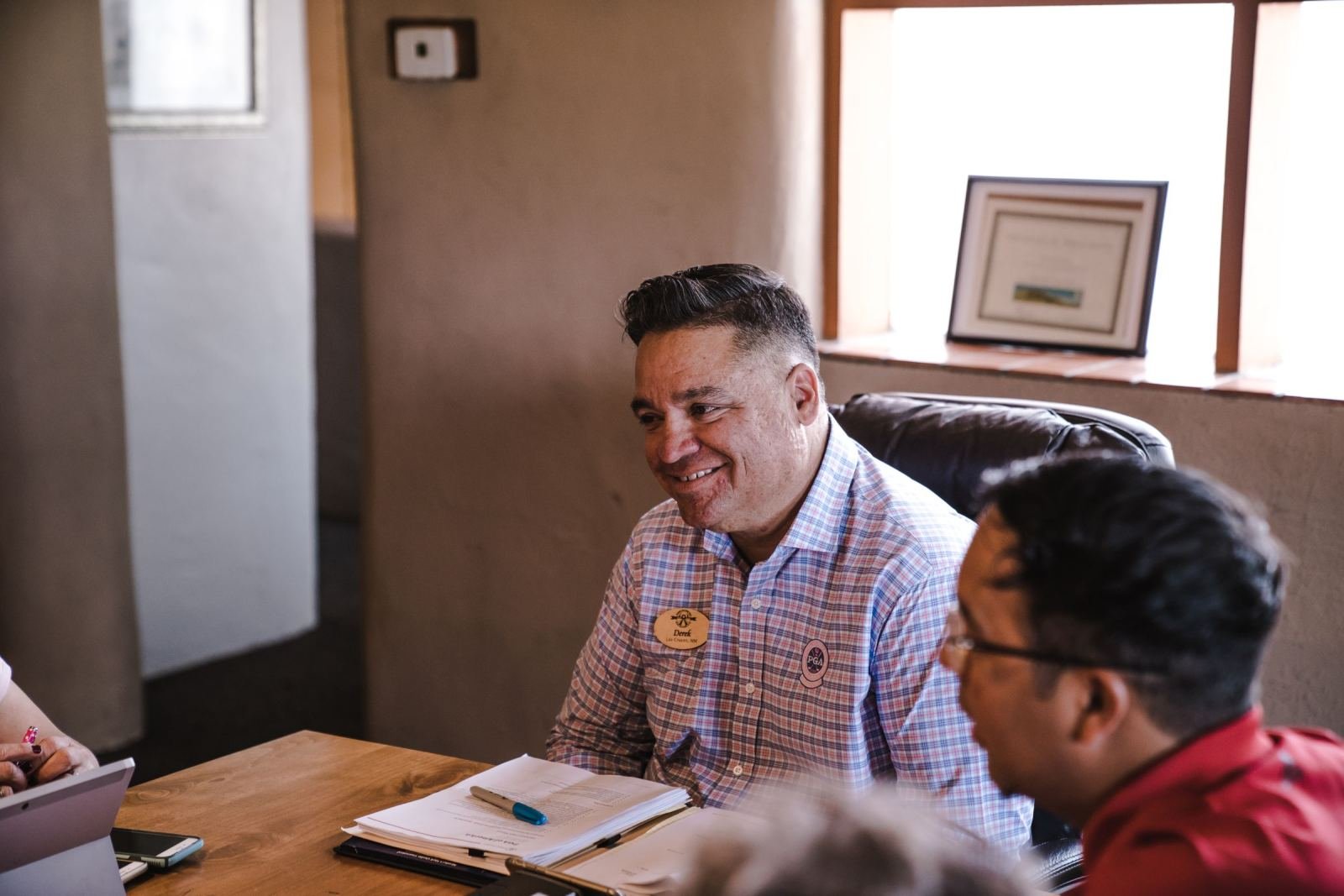 A man in a checkered shirt with a name tag talks with another man across a wooden table