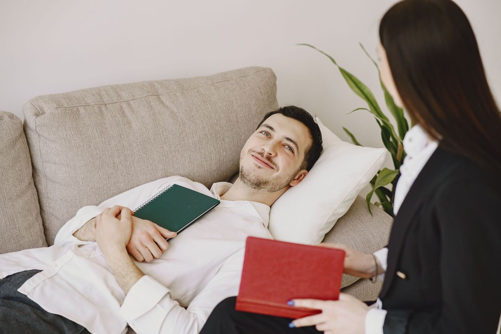 A man lies on a sofa with a notebook while a woman in a blazer talks to him