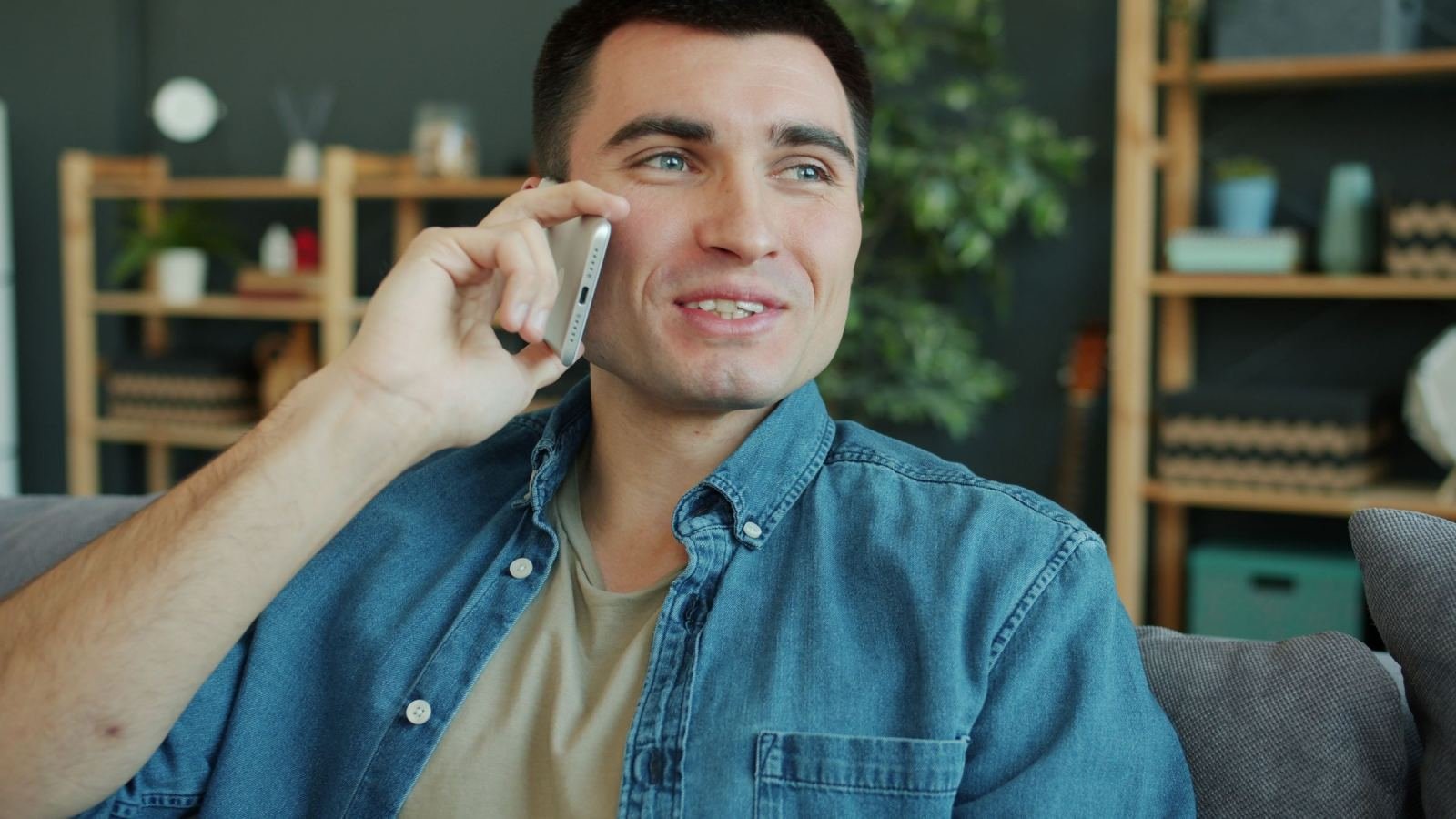 A man wearing a denim shirt sits on a couch and talks on his phone.