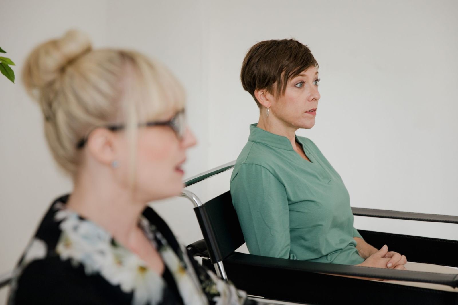 In a bright office, a woman wearing a green shirt talks to another woman.