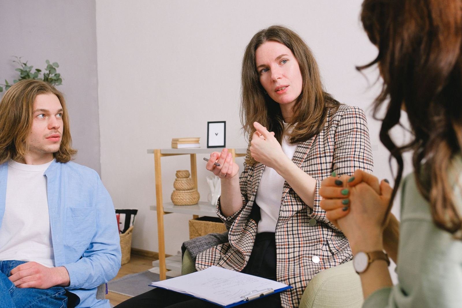 A woman in a plaid blazer speaks and gestures toward two people in a bright room