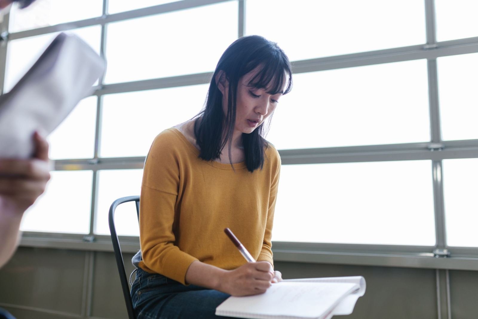 A woman in a yellow shirt writes in a notebook at a table