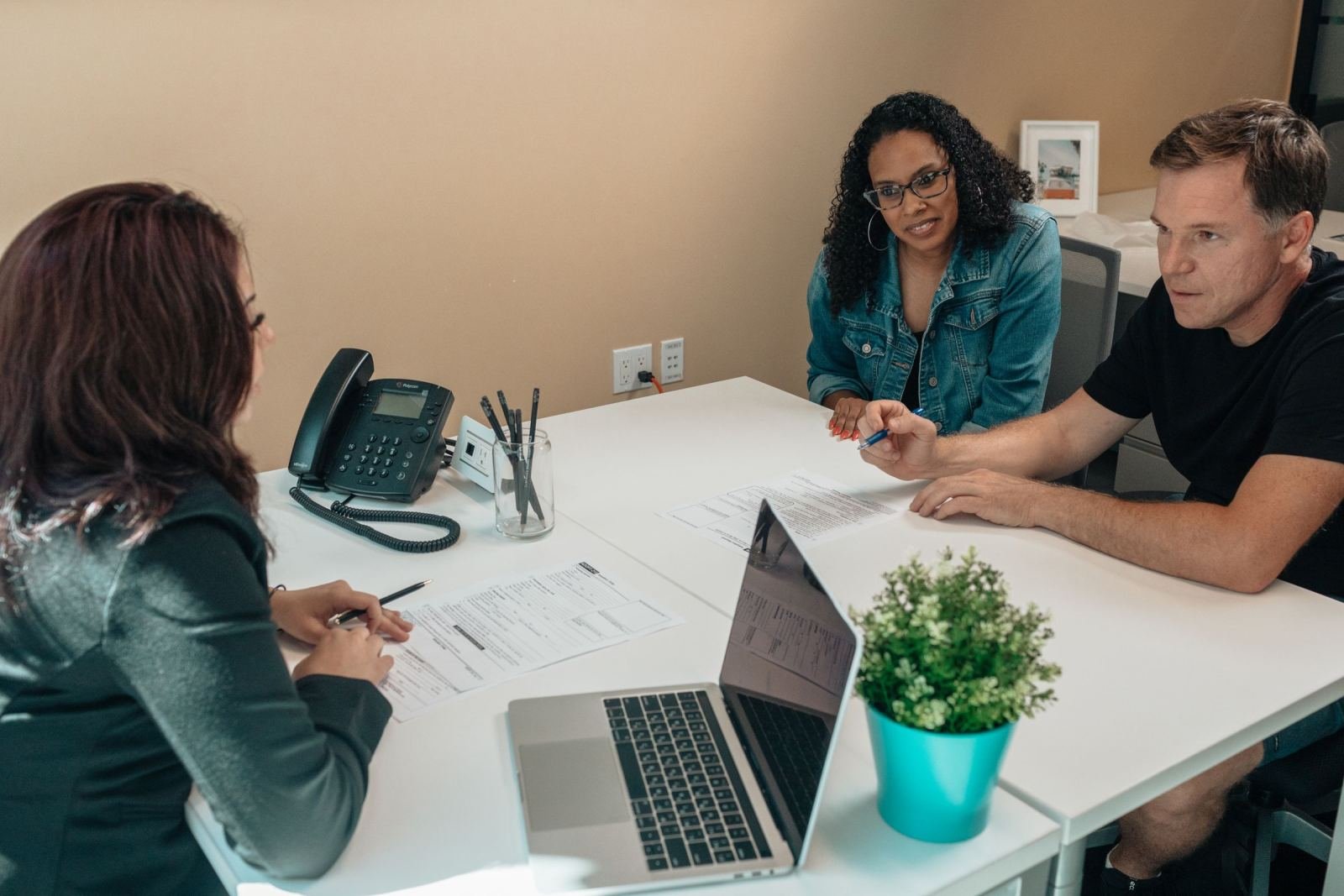 Three people sit around a white desk discussing documents and using a laptop