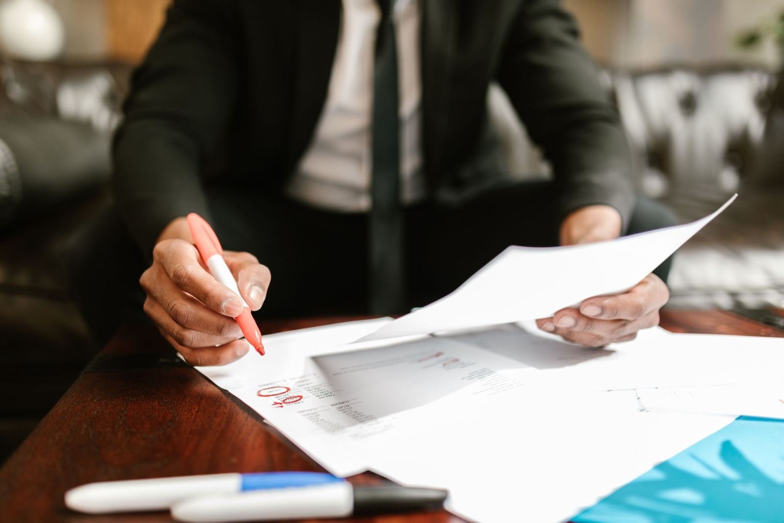 A person in a suit reviews documents with a red marker at a wooden table
