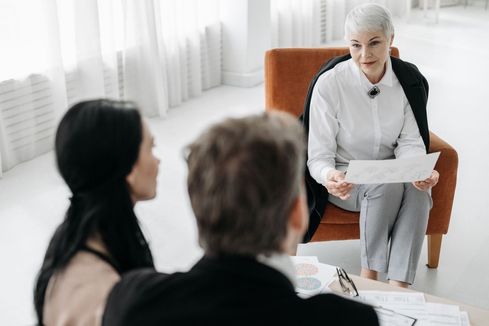 A professional woman in an orange chair speaks with two people while holding a document
