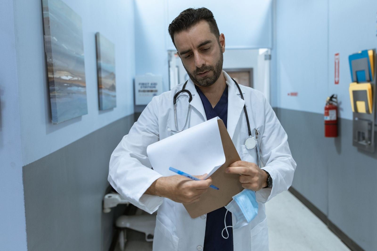 A doctor in a white coat looks at a clipboard in a hospital hallway
