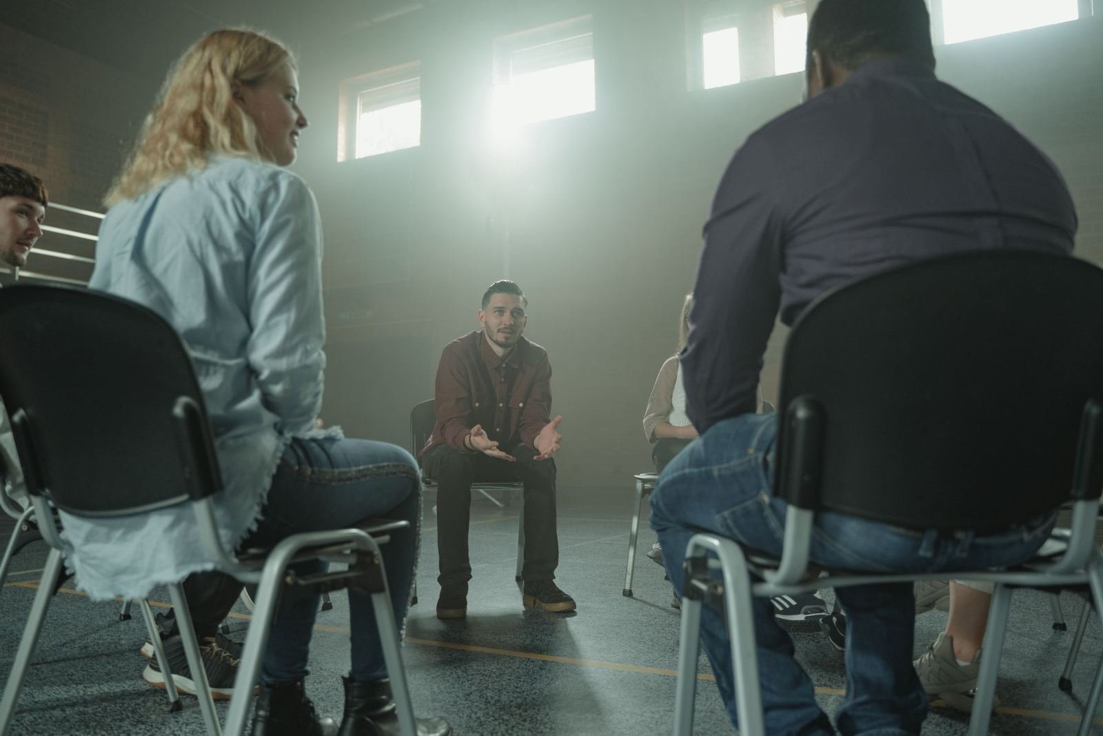 A group of people sit in a circle of chairs while a man in a red shirt speaks