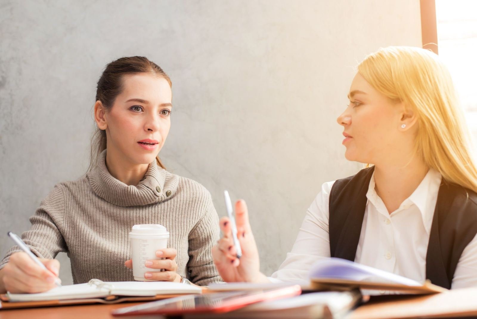 Two women sit at a table with coffee and notebooks.