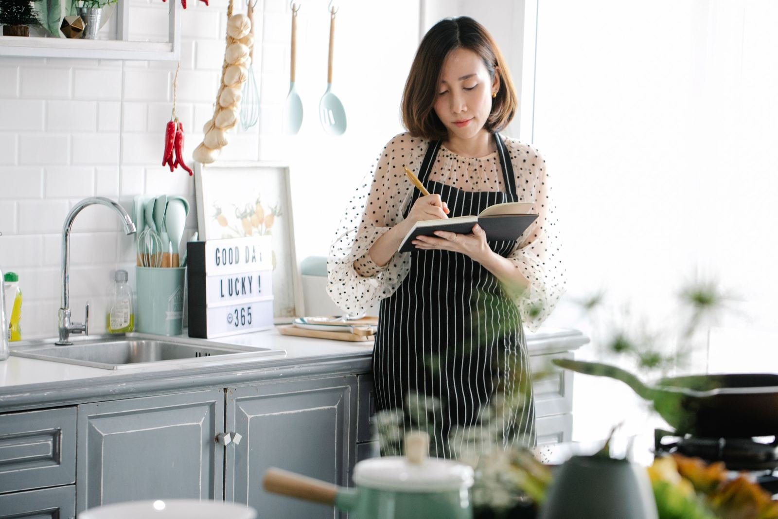 A woman in a striped apron writes in a notebook in a bright kitchen
