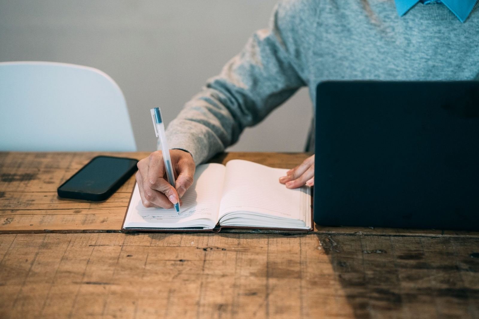 A person writing in a notebook at a wooden table with a laptop and phone