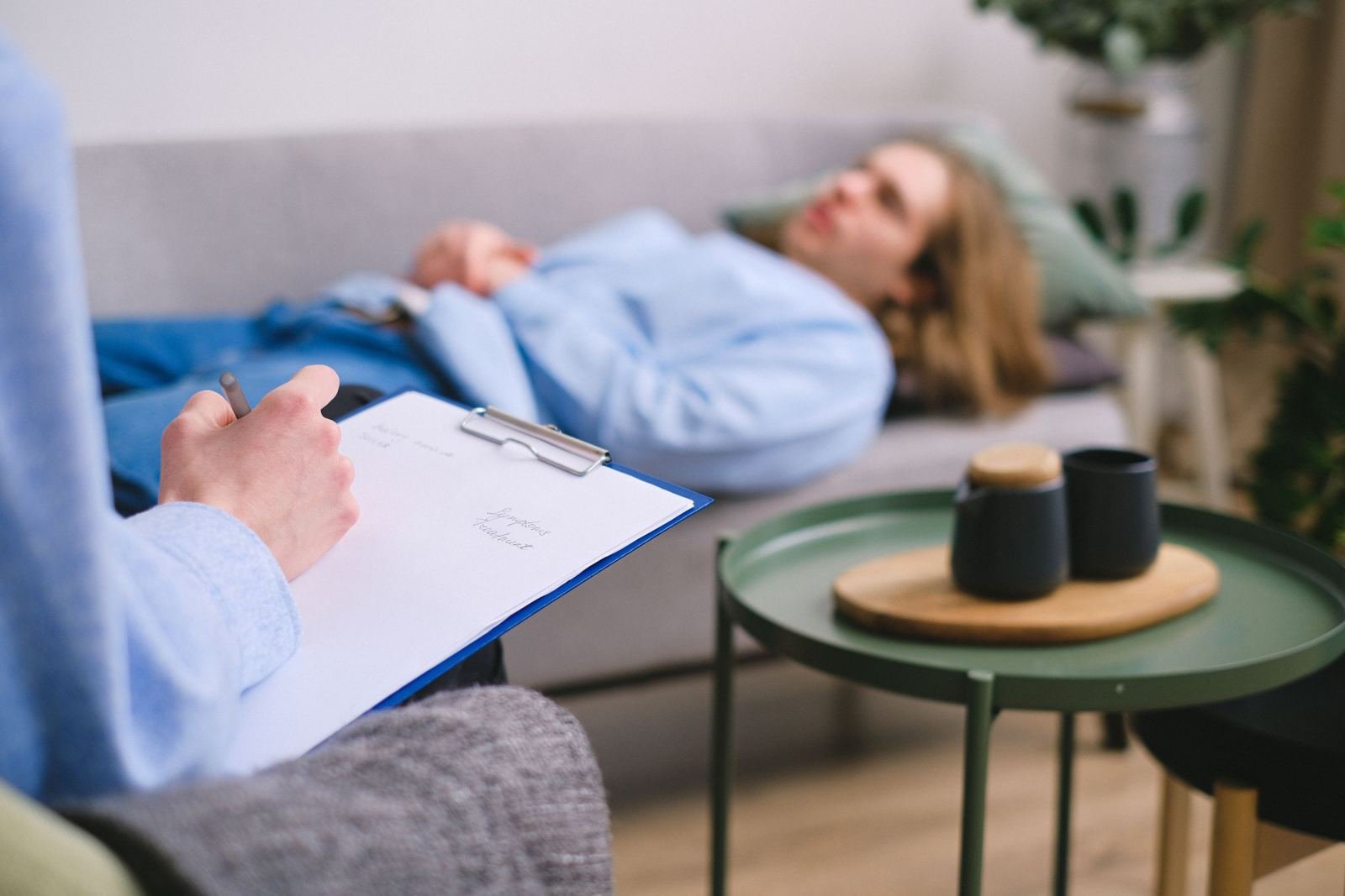 A professional writes on a clipboard while a person lies on a sofa in the background