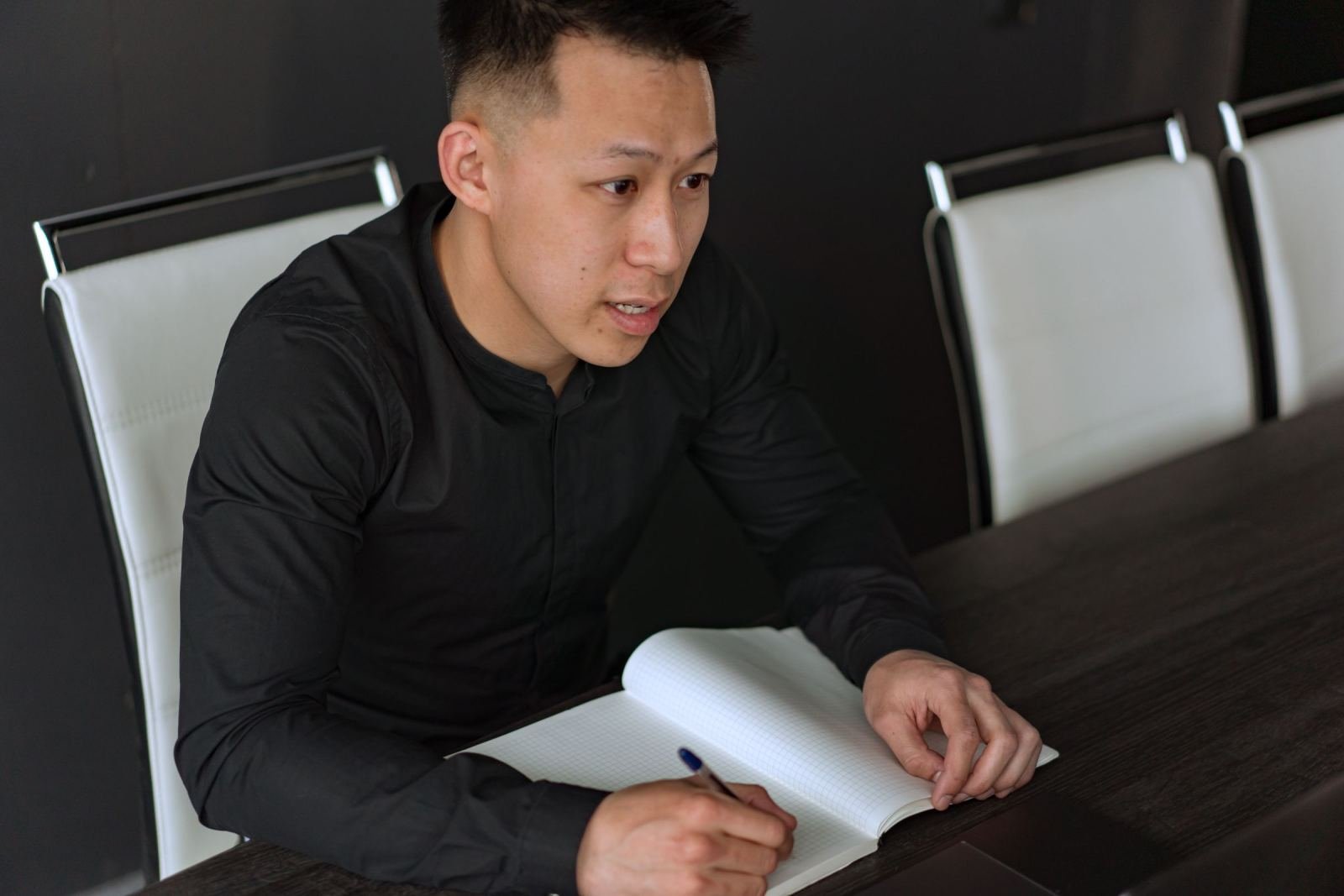 A man in a black shirt writes in a notebook while sitting at a conference table