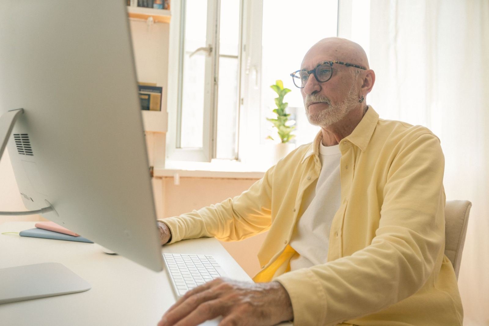 An older man with glasses looks at a desktop computer monitor at a white desk
