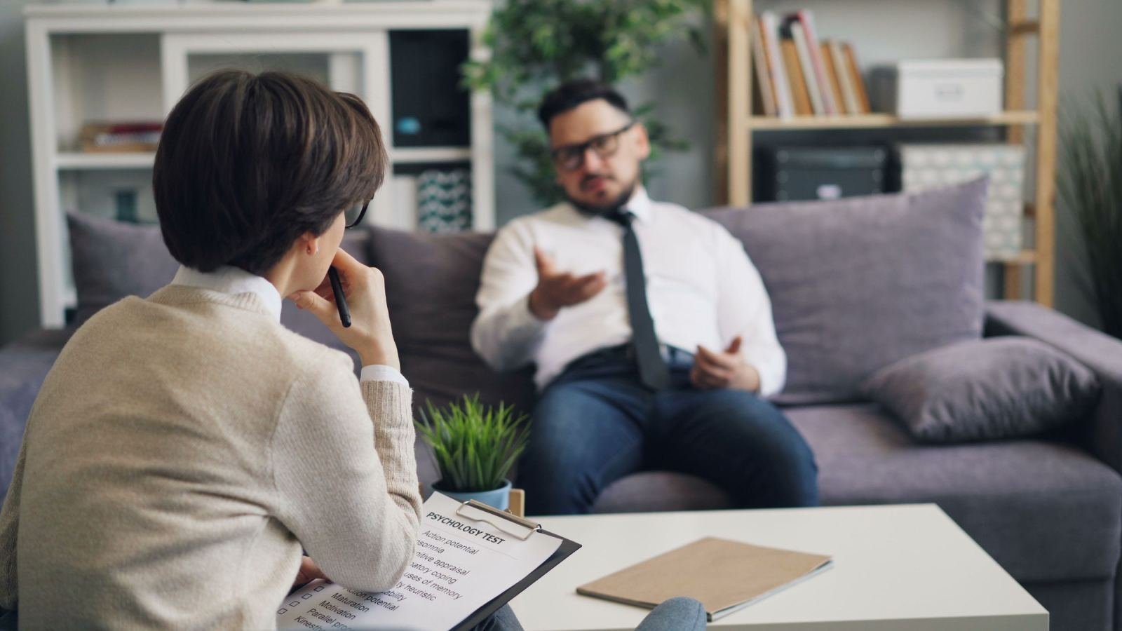A man in a white shirt talks to a clinician holding a clipboard in a bright office