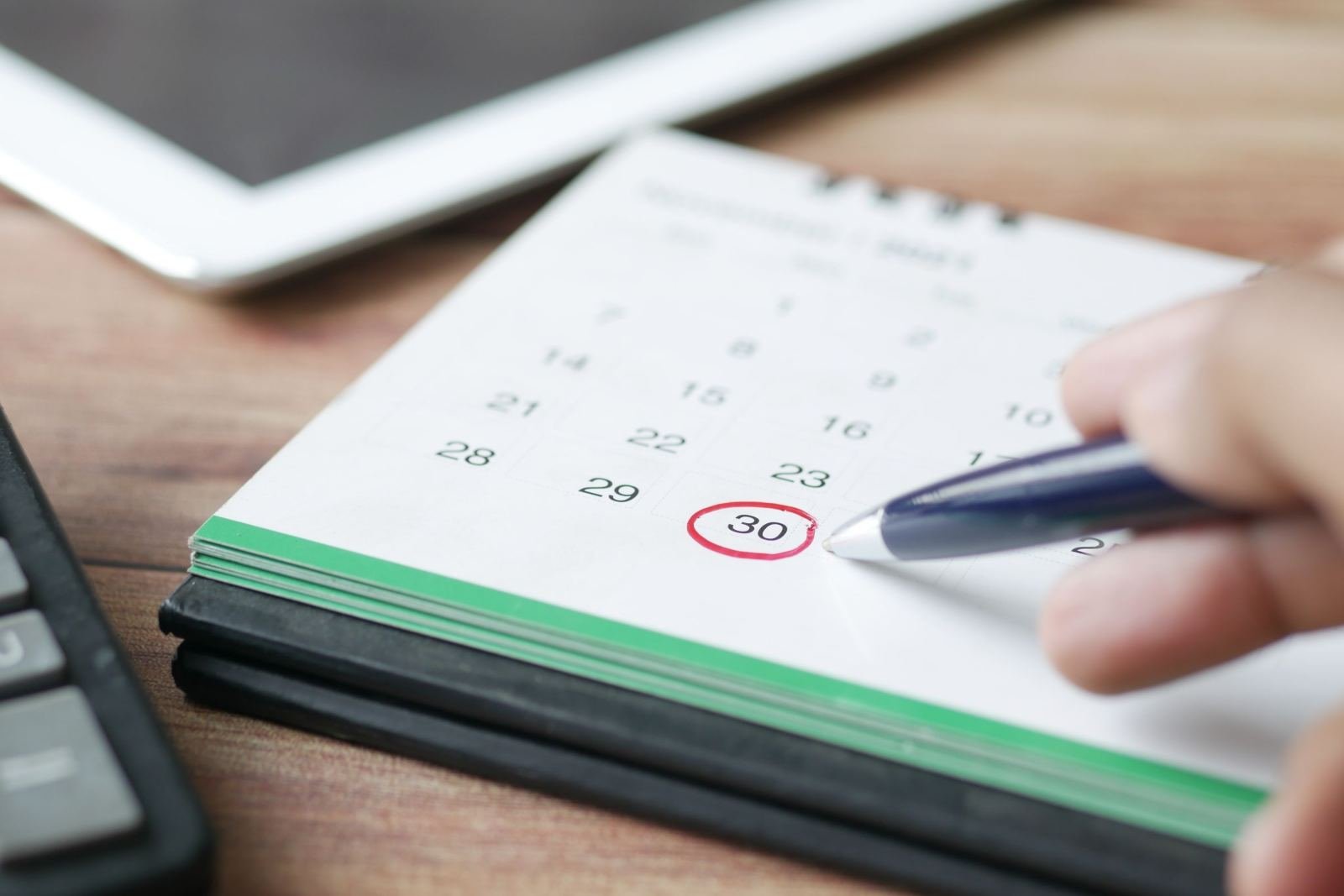 A hand uses a blue pen to circle the 30th on a desk calendar