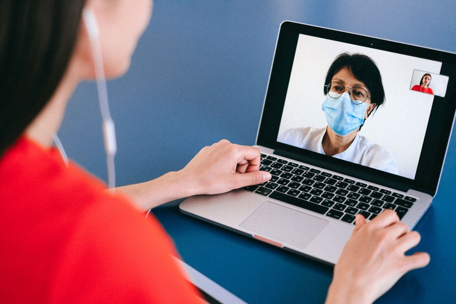 A person in a red shirt talks to a masked healthcare provider on a laptop video call.