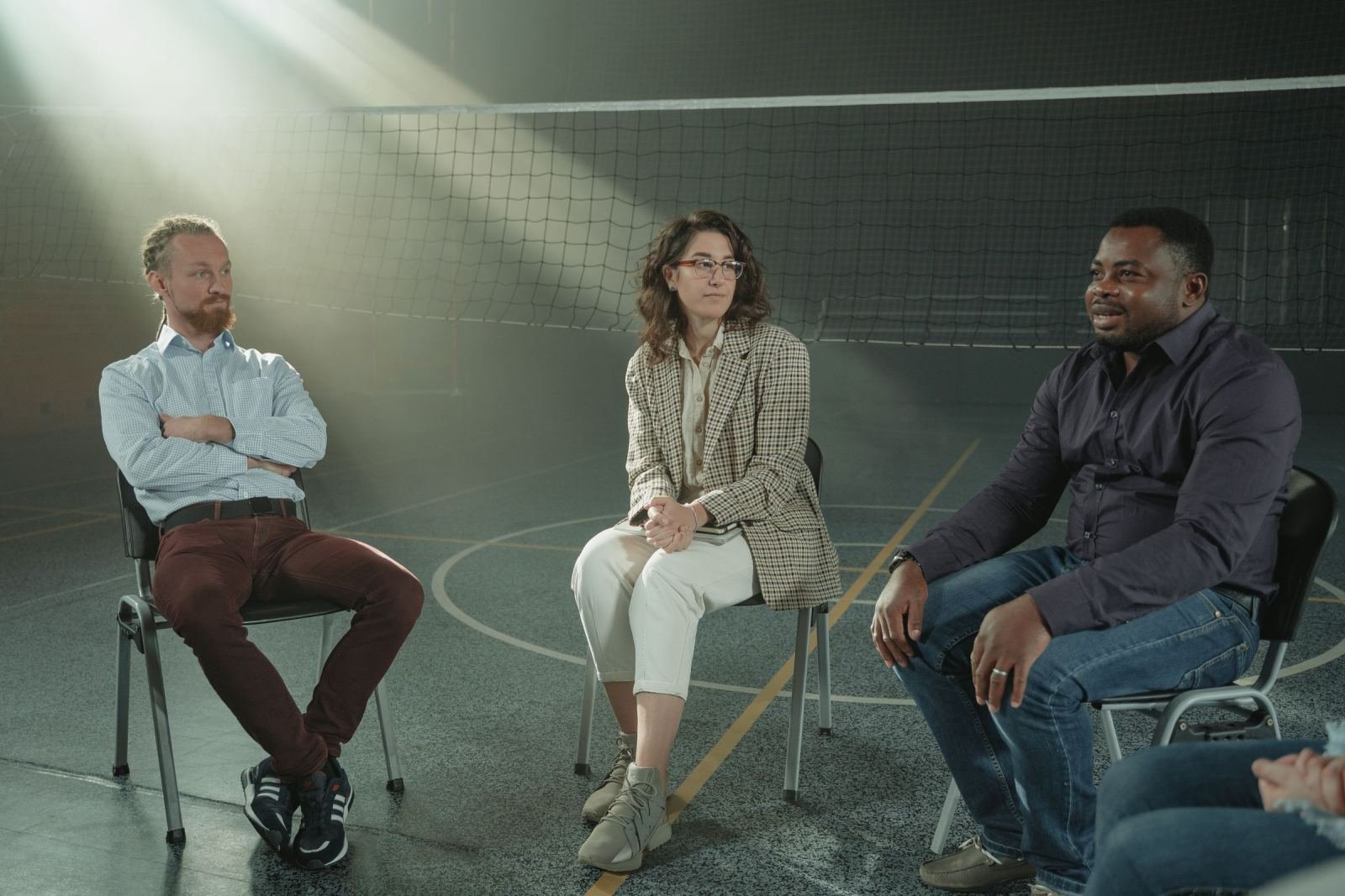Three people sit in a circle and talk in a large room with a volleyball net
