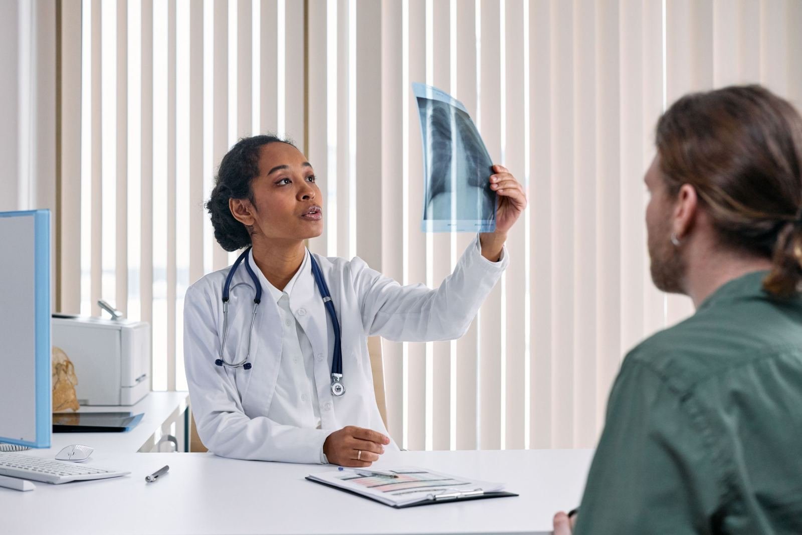 A clinician in a white coat shows an X-ray to a patient in a bright office