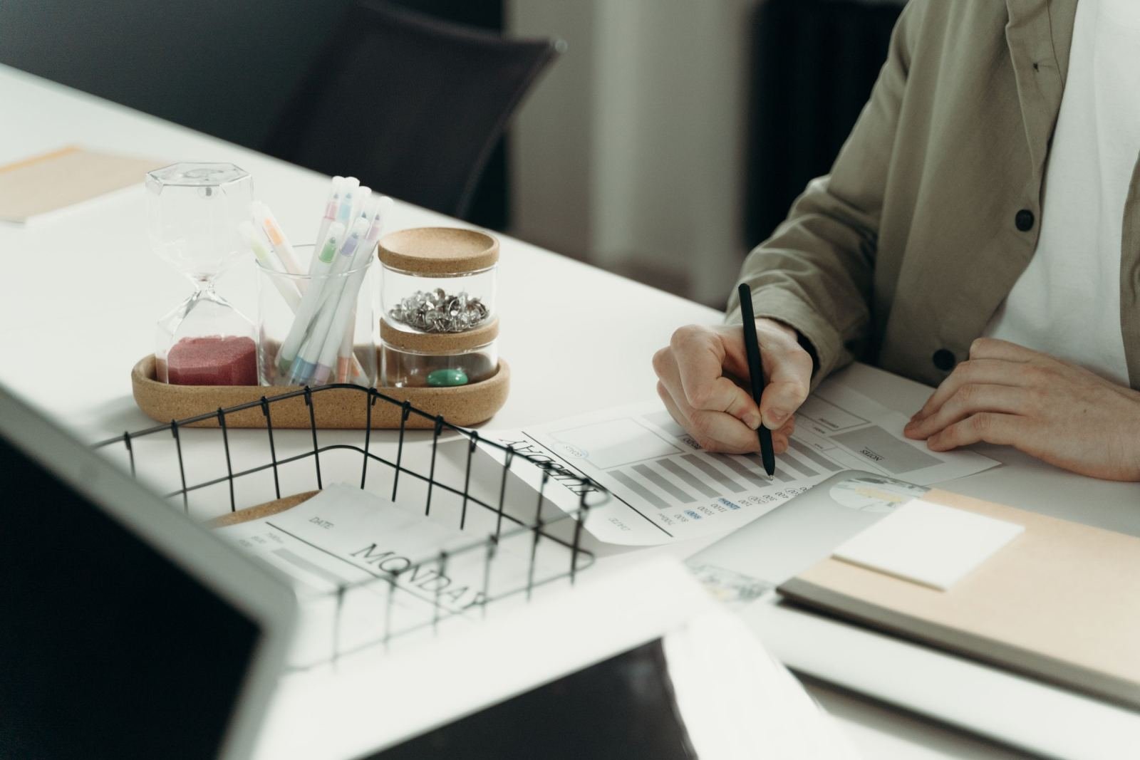 A person in a tan blazer writes on a document at a white desk with office supplies