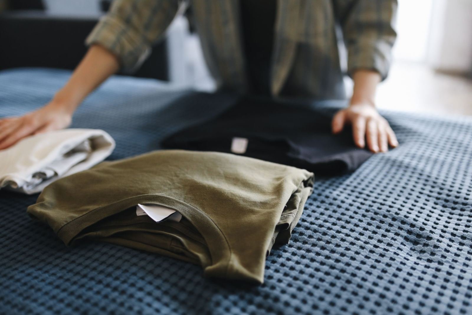 A person arranges folded white, green, and black shirts on a blue bedspread