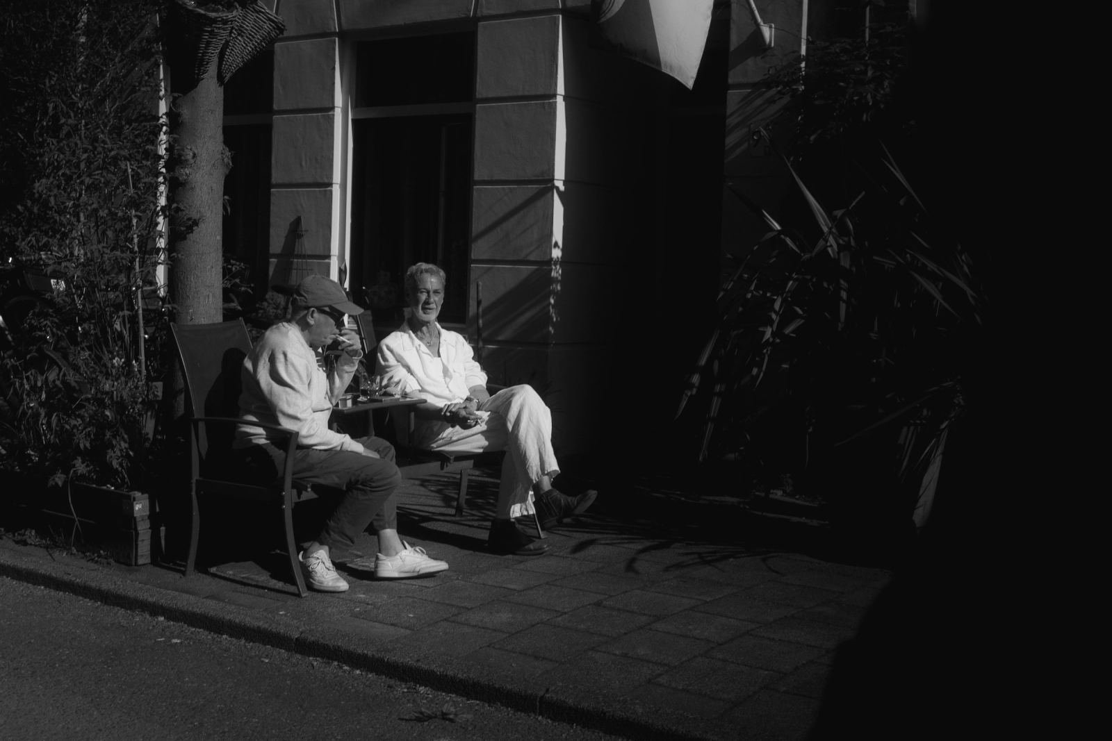 Two people sit and talk at a small outdoor table in the shade