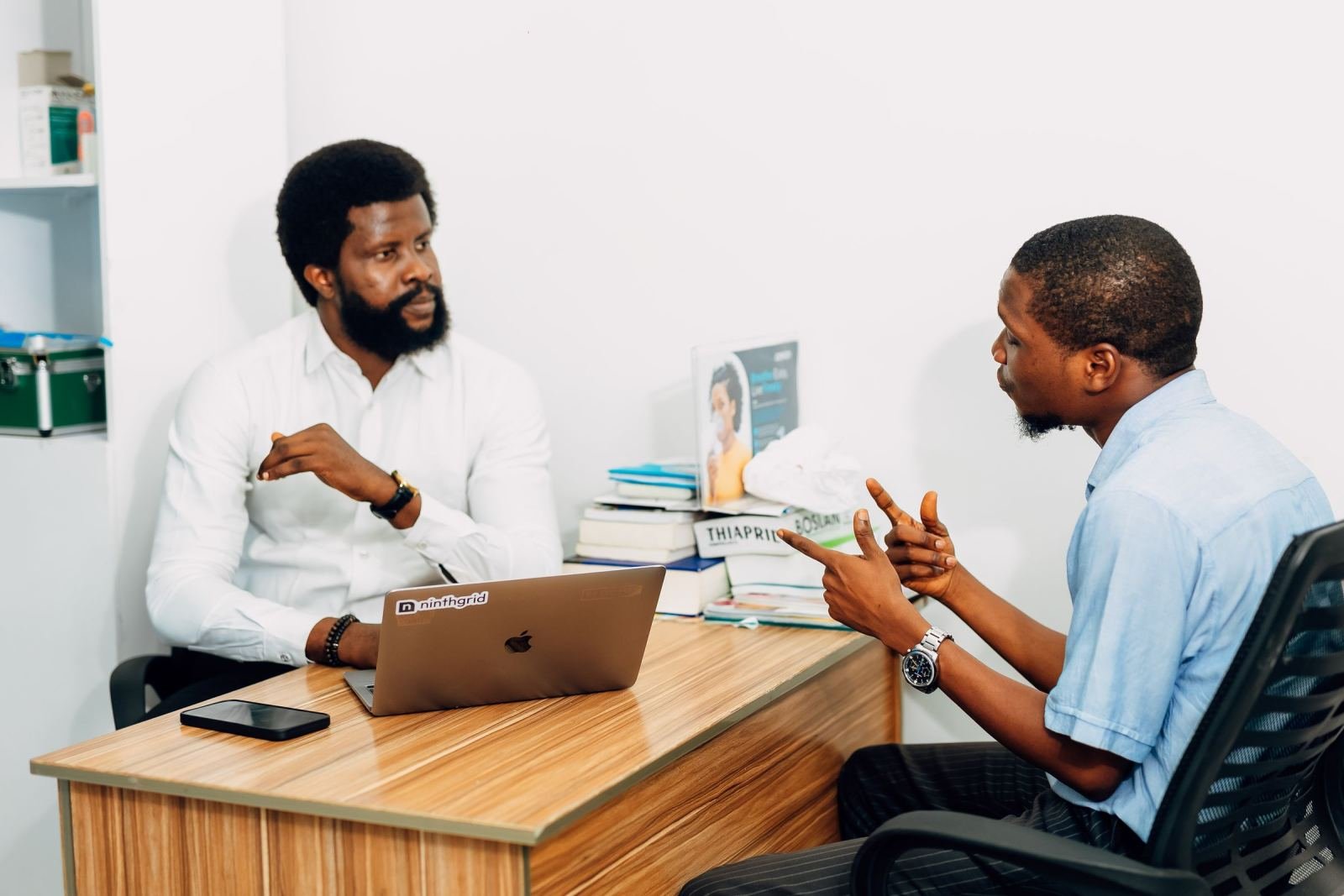 Two men talking across a desk with a laptop and books in a bright room
