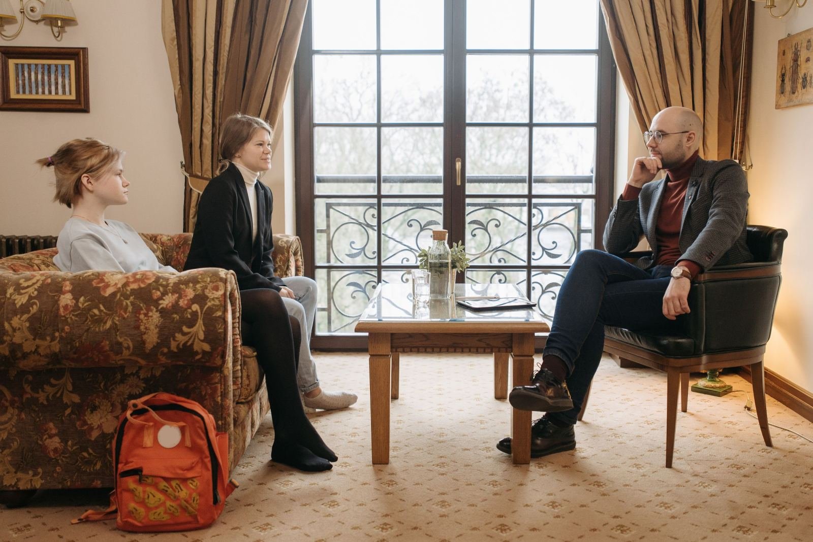 A man in a blazer listens to two women in a living room setting