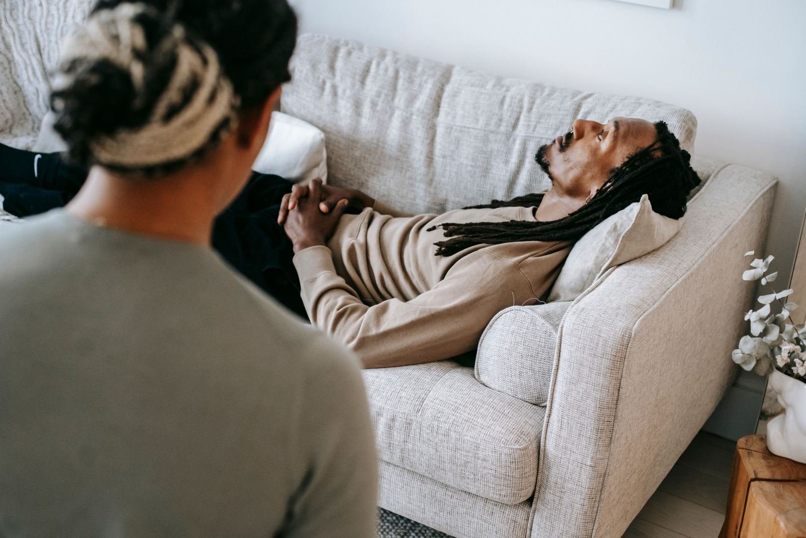 A man with dreadlocks lies on a couch, speaking with someone else.