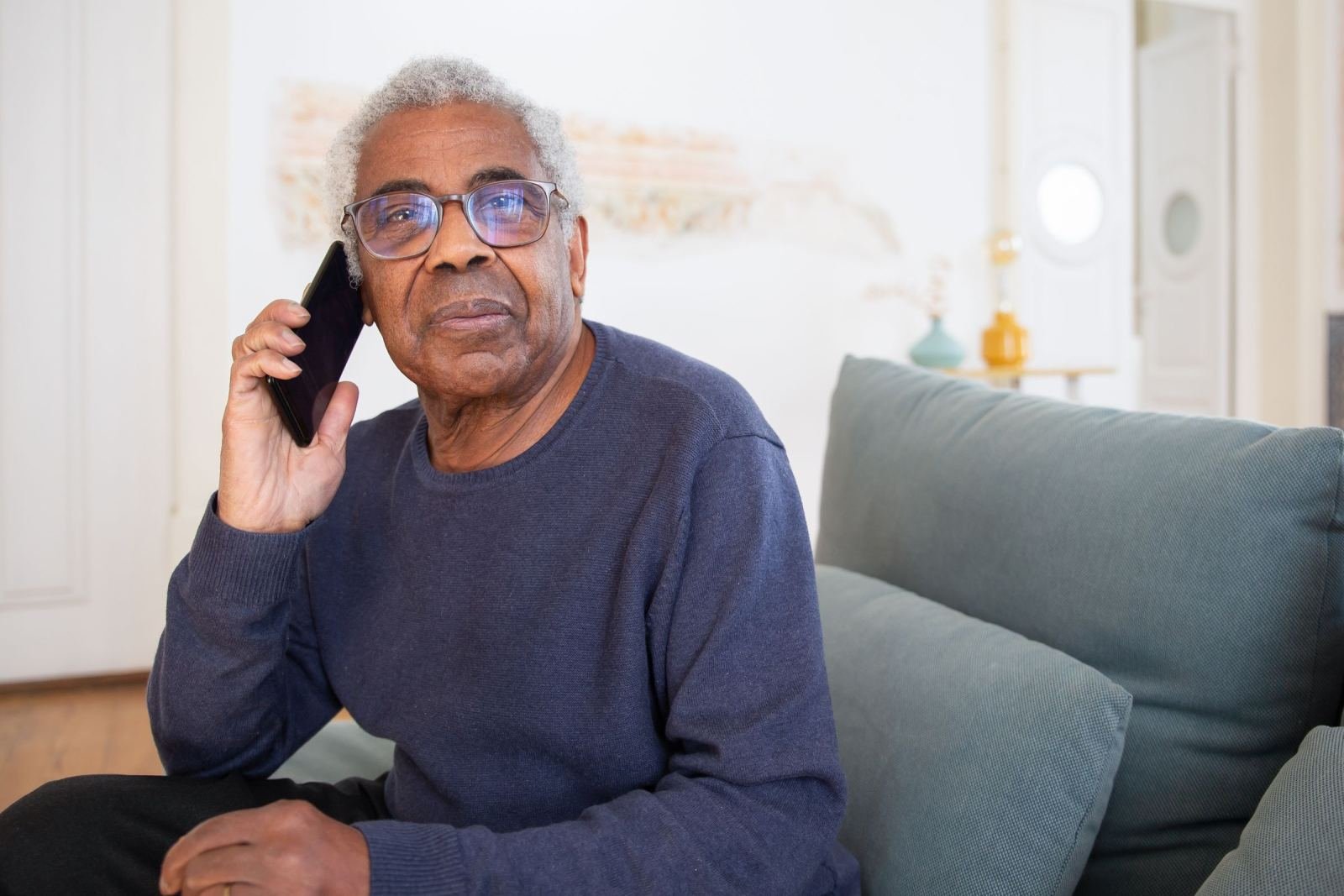 An older man with glasses talks on a smartphone while sitting on a gray sofa