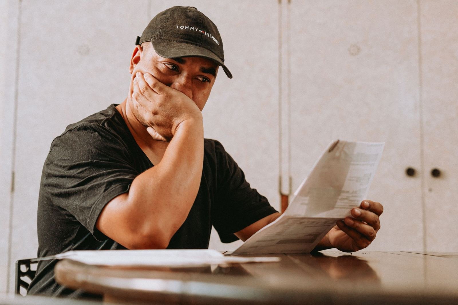 A man in a black cap reads a document while resting his chin on his hand