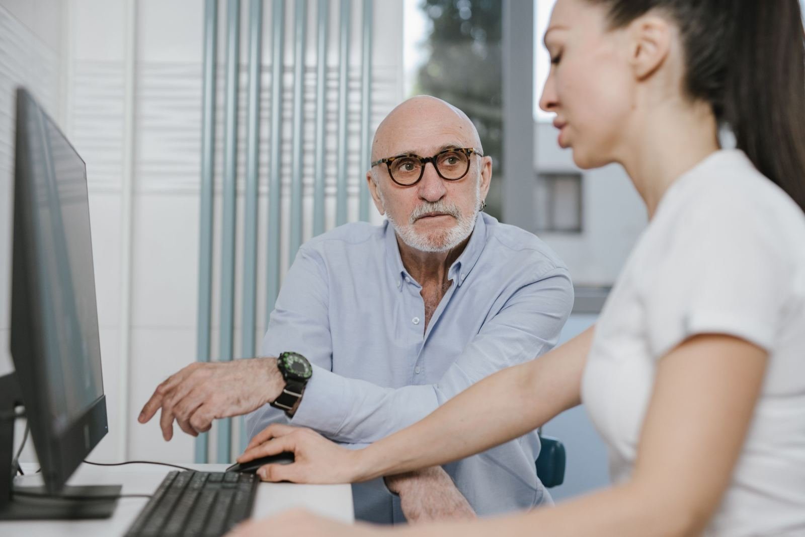 An older man and a woman look at a computer screen together in a bright office