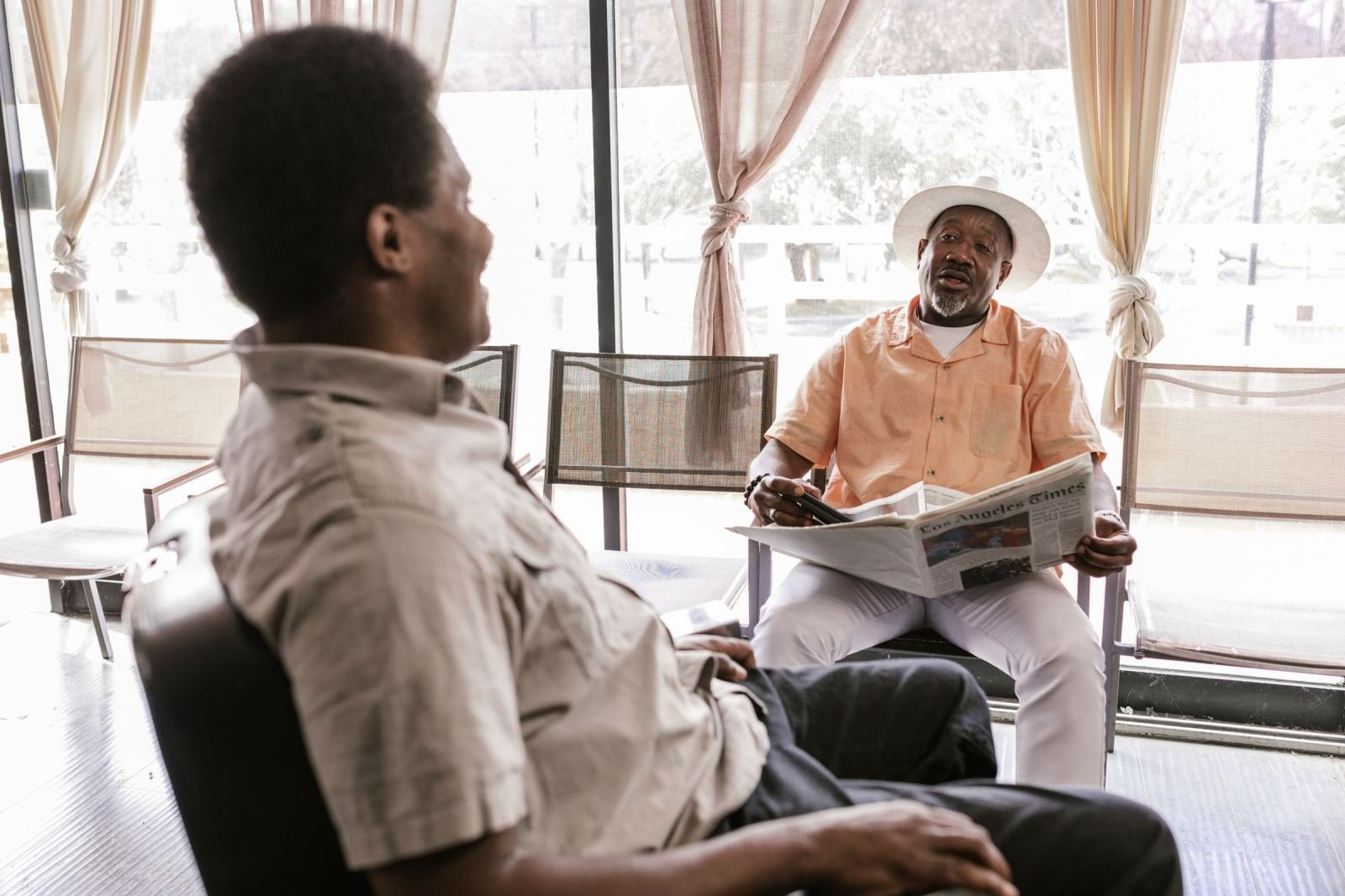 Two men talking in a bright room, one wearing a white hat and holding a newspaper