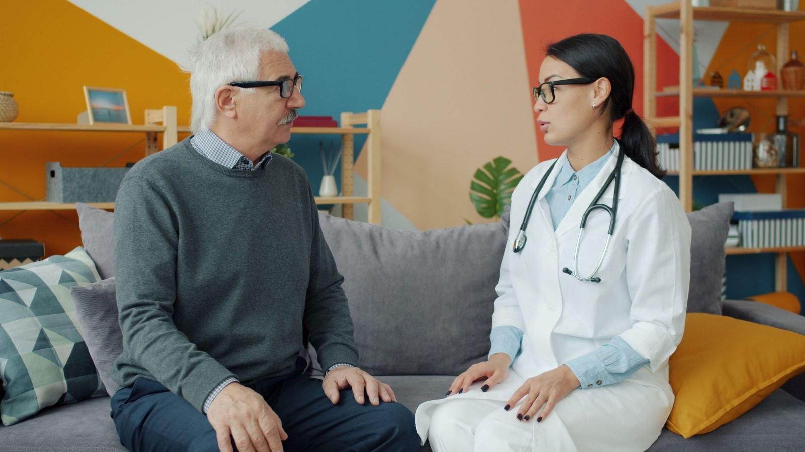 A doctor in a white coat sits on a sofa and talks to an older man.