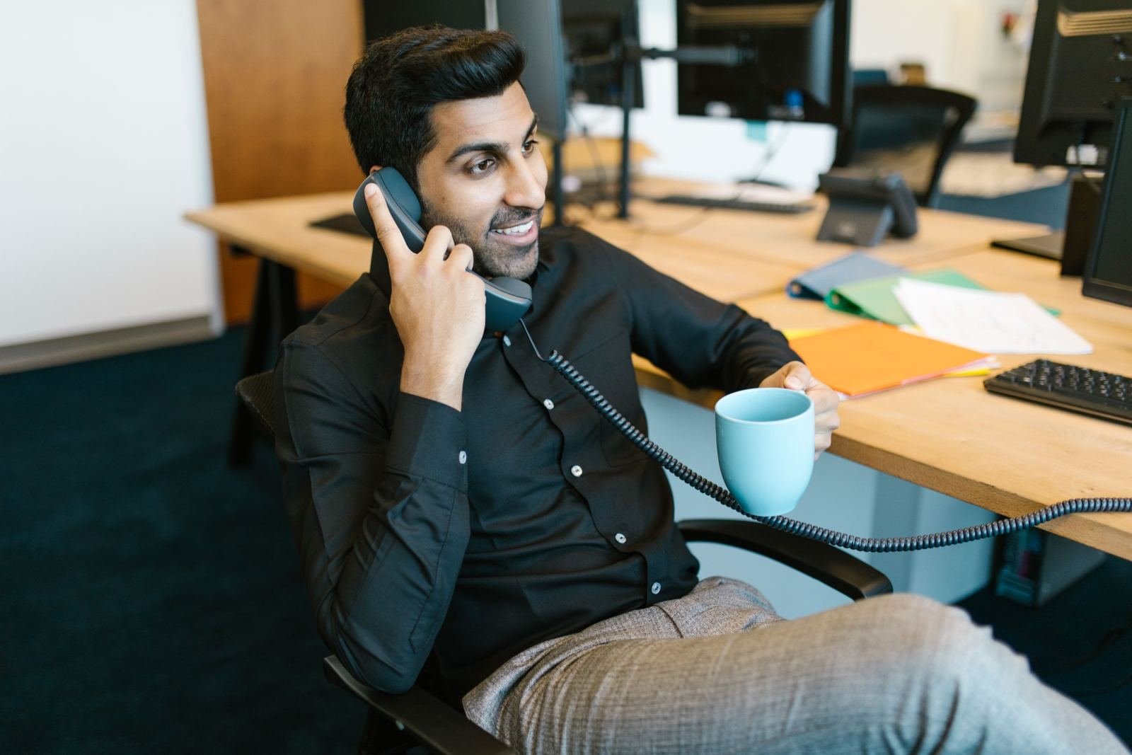 A man in a black shirt talks on a desk phone while holding a blue mug