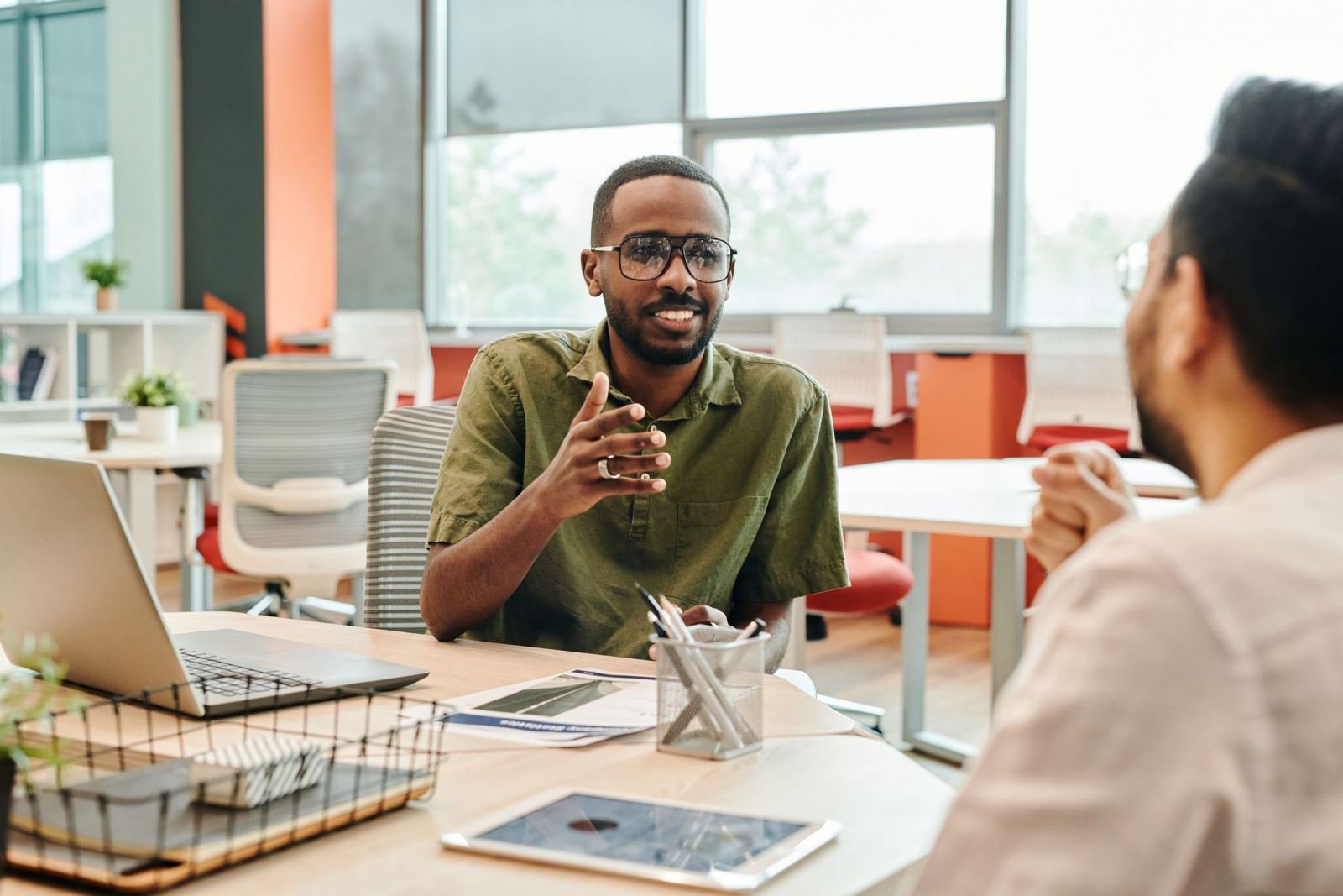 A man wearing a green shirt sits across from someone in an office. He is talking and using his hands.