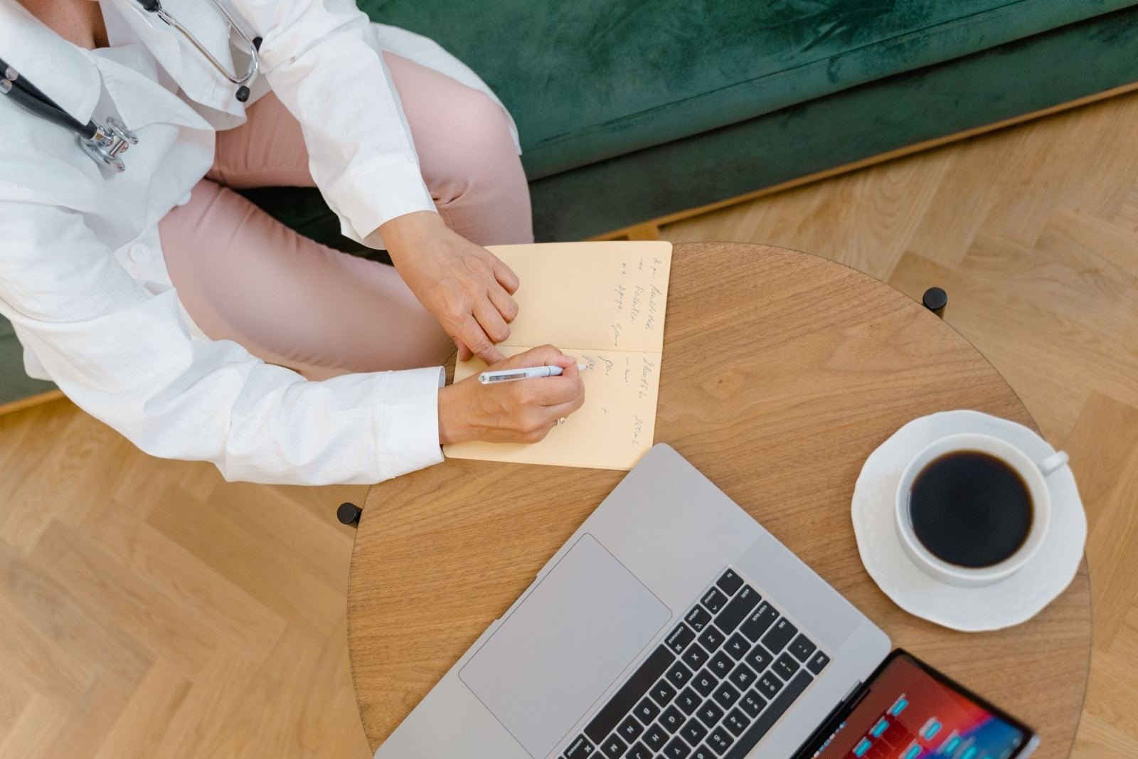 A clinician in a white coat writing in a notebook next to a laptop and coffee