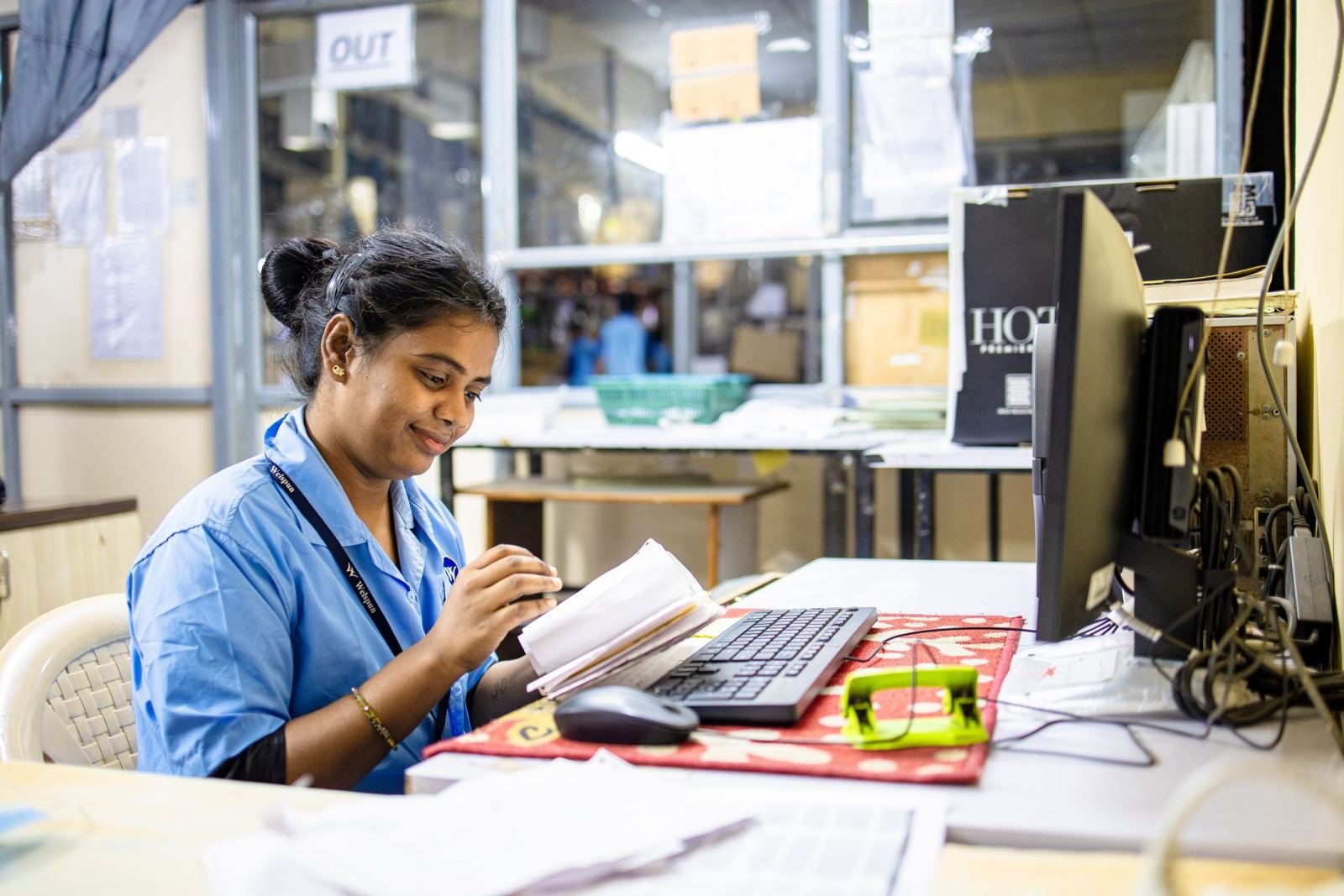 A woman in a blue uniform looks at a notebook while sitting at a computer desk