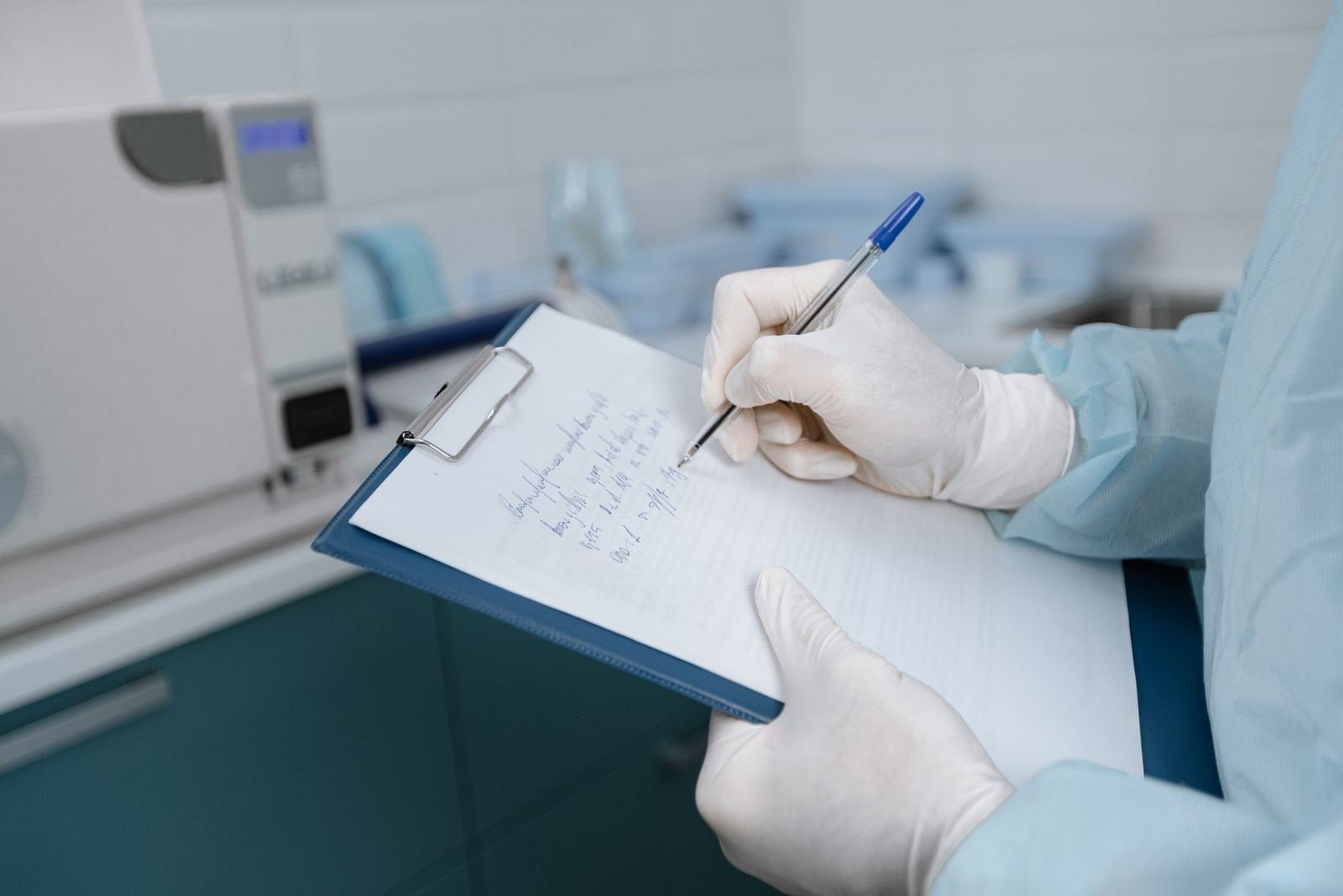 A clinician in blue scrubs and white gloves writing notes on a clipboard