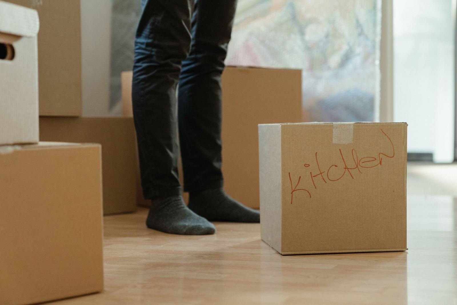 A person stands among cardboard boxes, one labeled Kitchen, on a wooden floor