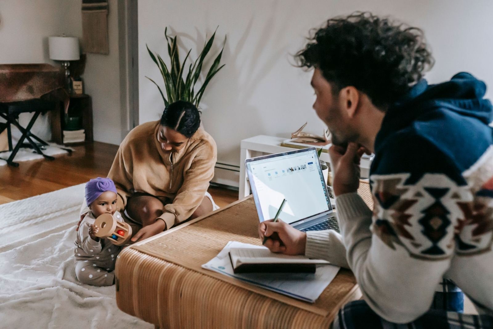 A man takes notes in a book beside his laptop. A woman and child are sitting close by.