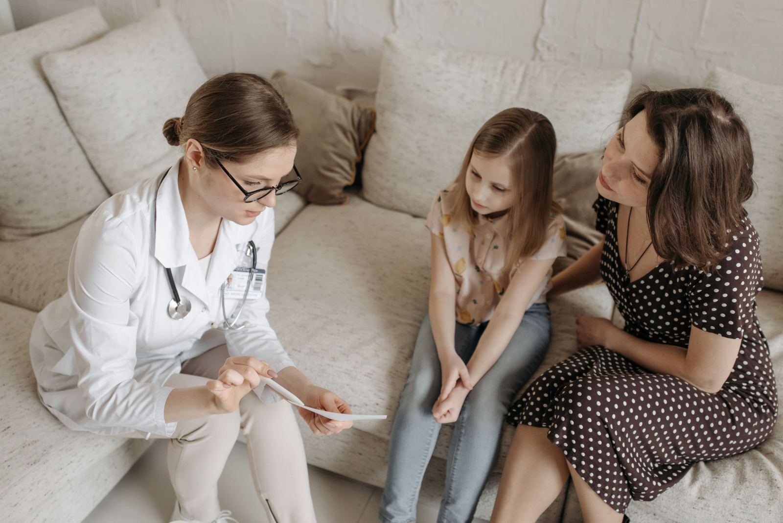 A clinician in a white coat discusses a document with a woman and a young girl on a sofa