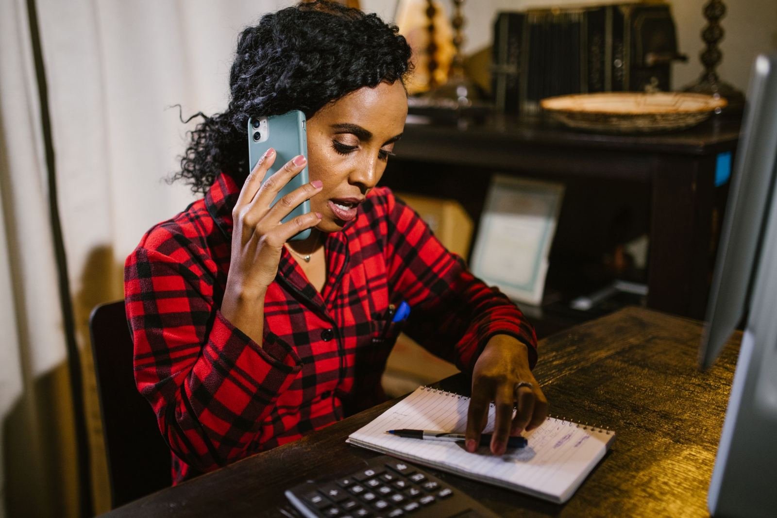 A woman uses a phone and writes notes at her desk.