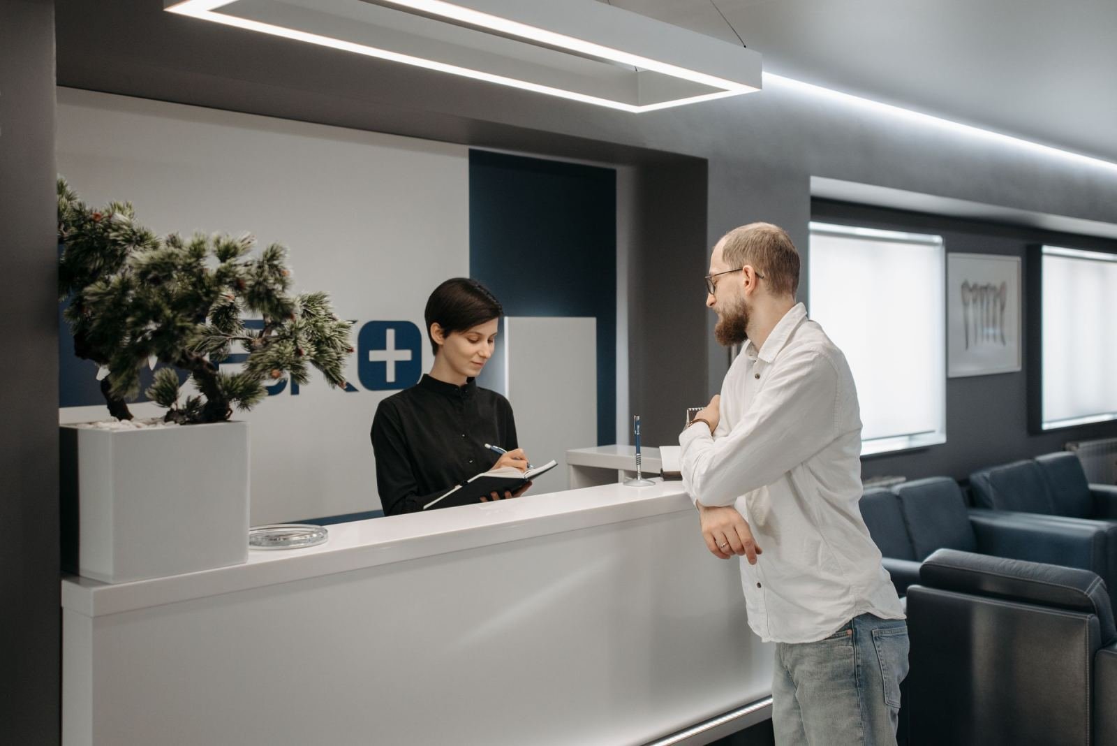 A woman at the front desk takes notes in a book while speaking with a man.