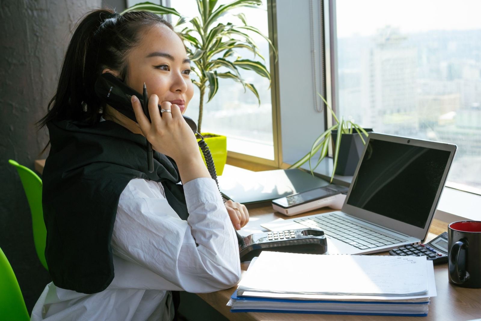 A woman on a corded phone sits at a desk with a laptop and papers