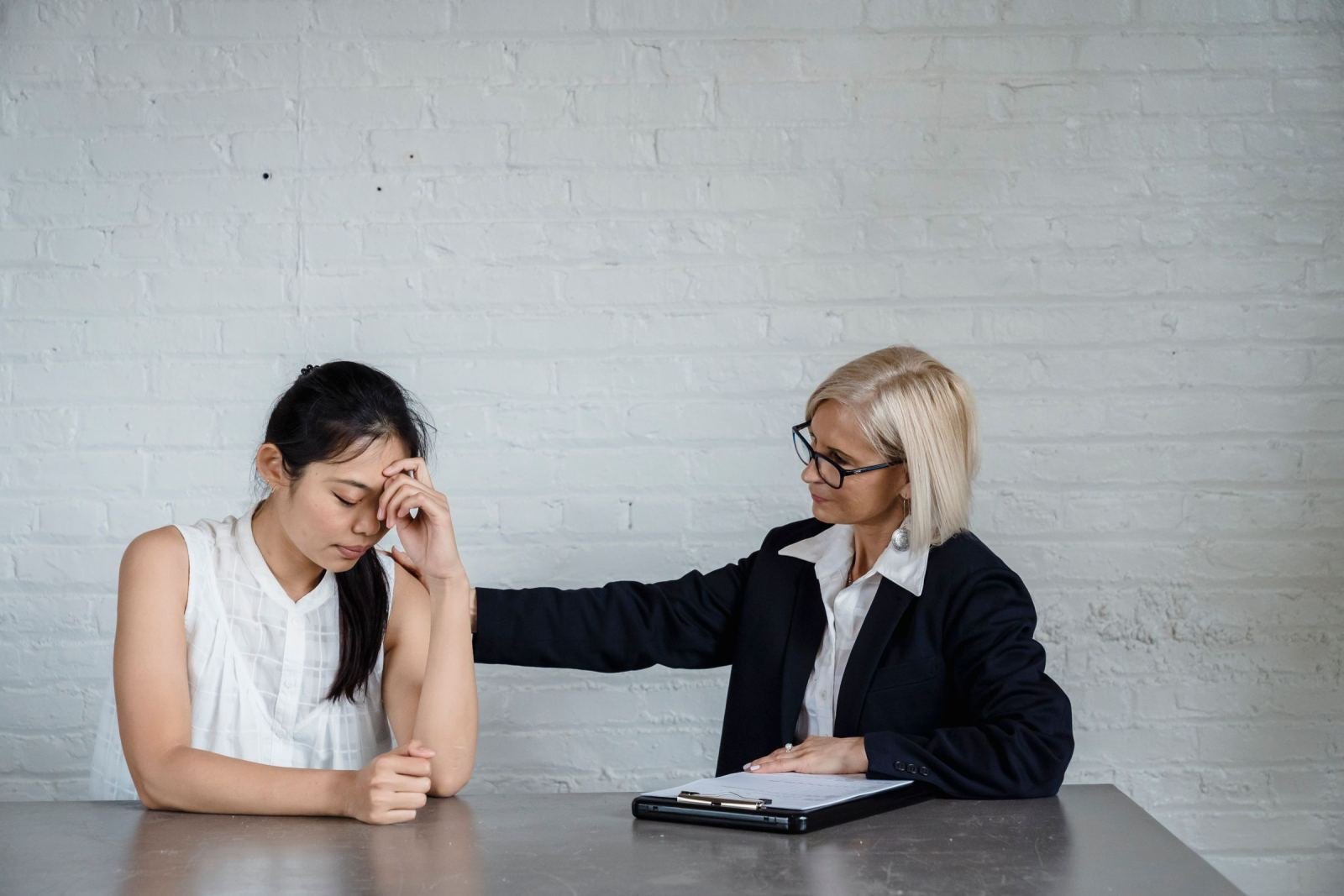 A woman with her hand on her forehead sits with a professional woman who has a hand on her shoulder