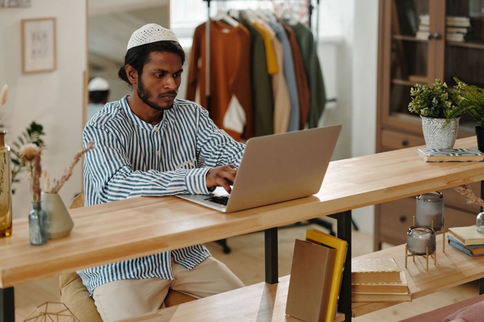 A man wearing a striped shirt and white skullcap sits at a wooden table using a laptop.