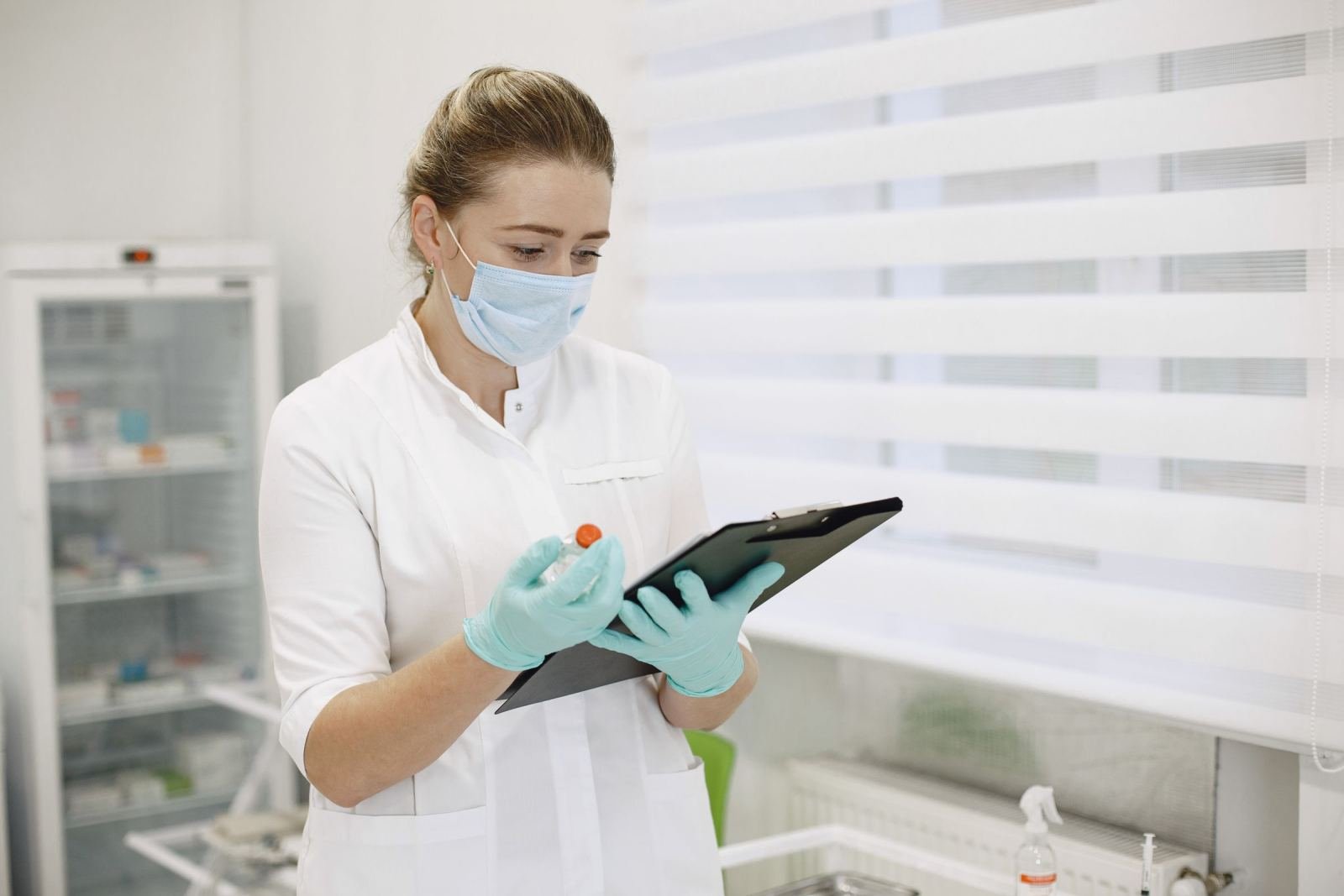 A medical professional in a mask and gloves holds a vial and a clipboard